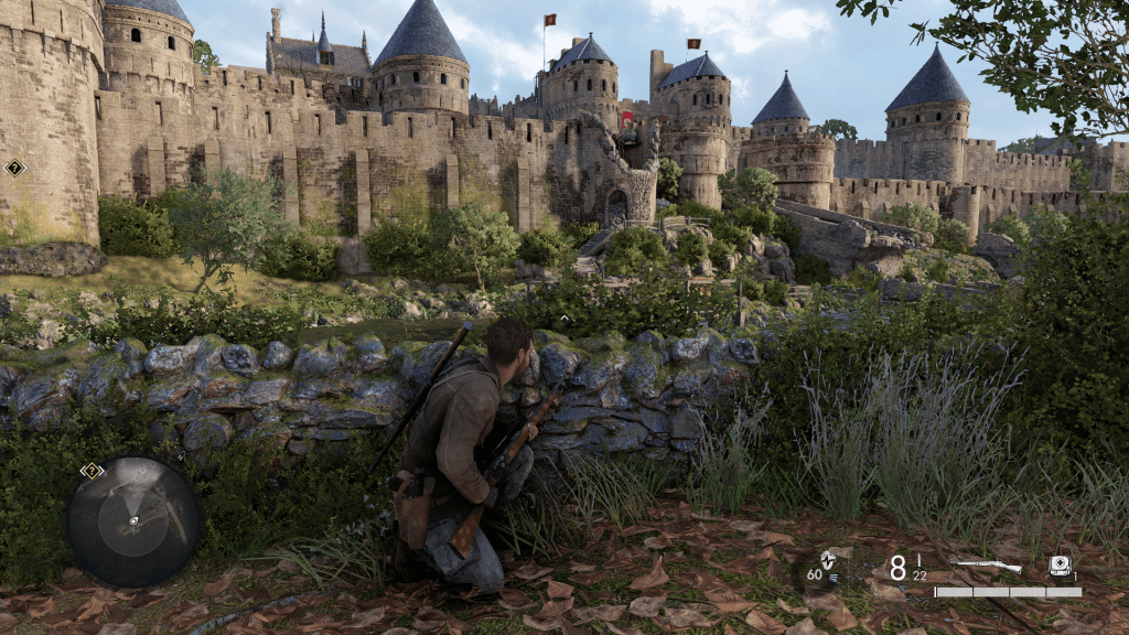 A sniper crouching behind a stone wall, observing a heavily fortified medieval castle occupied by enemy forces in Sniper Elite: Resistance. The environment is lush with greenery, and the castle has multiple towers with red flags.