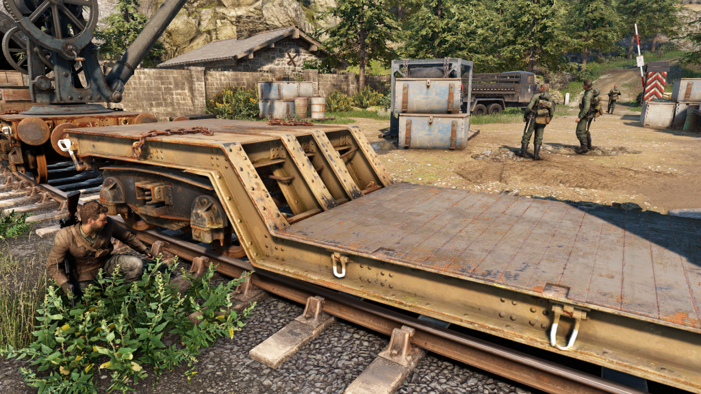 A sniper crouches behind a train cart, hiding from a group of enemy soldiers in a military rail yard in Sniper Elite: Resistance. The environment features industrial structures, supply crates, and a truck in the background.