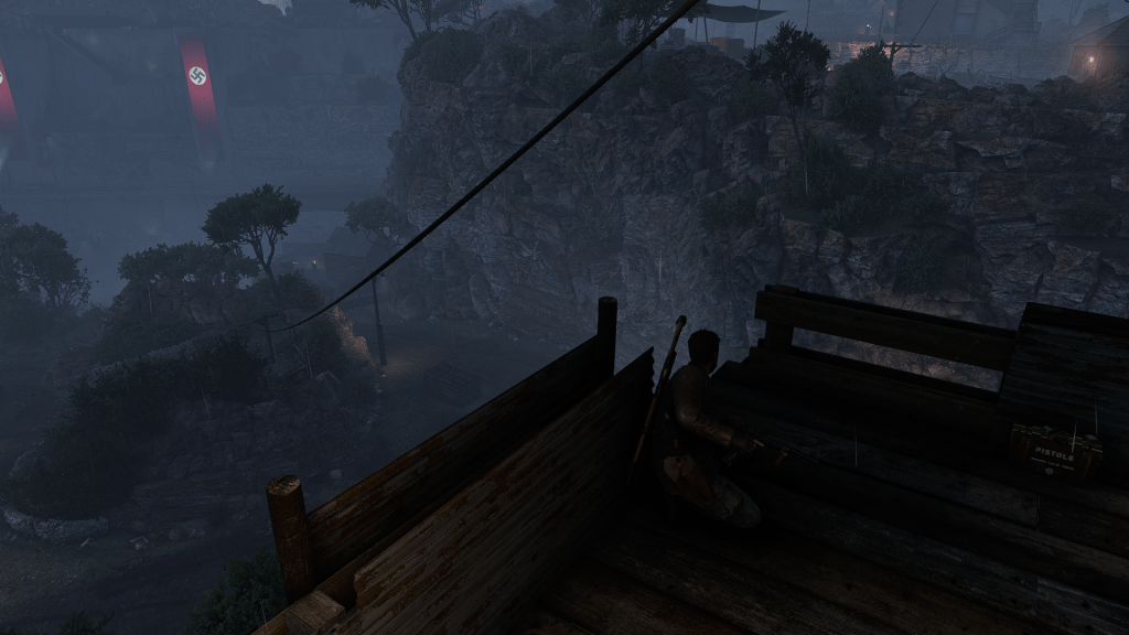 A sniper crouches in a wooden platform at night, overlooking a Nazi-controlled facility built into a rocky canyon. A zipline extends into the distance, and rain falls over the dark, war-torn landscape.